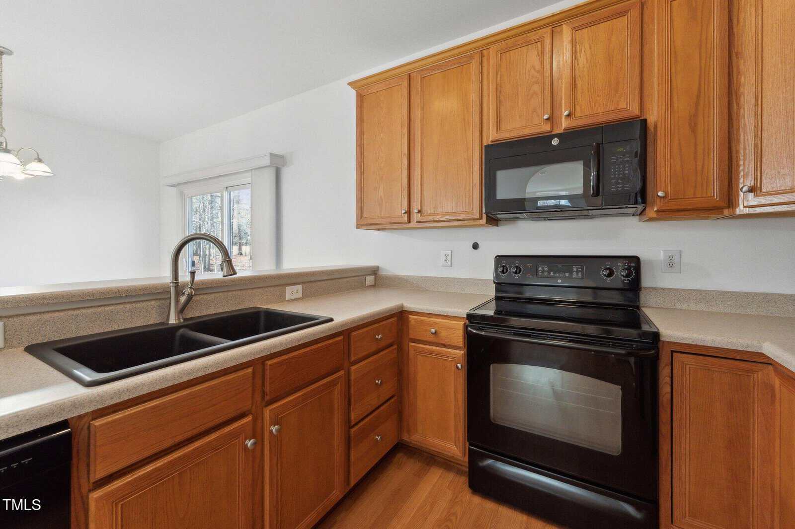 A kitchen with wooden cabinets, black electric stove and oven, black microwave, double black sink with a silver faucet, and beige countertops. A window with vertical blinds is visible in the background.