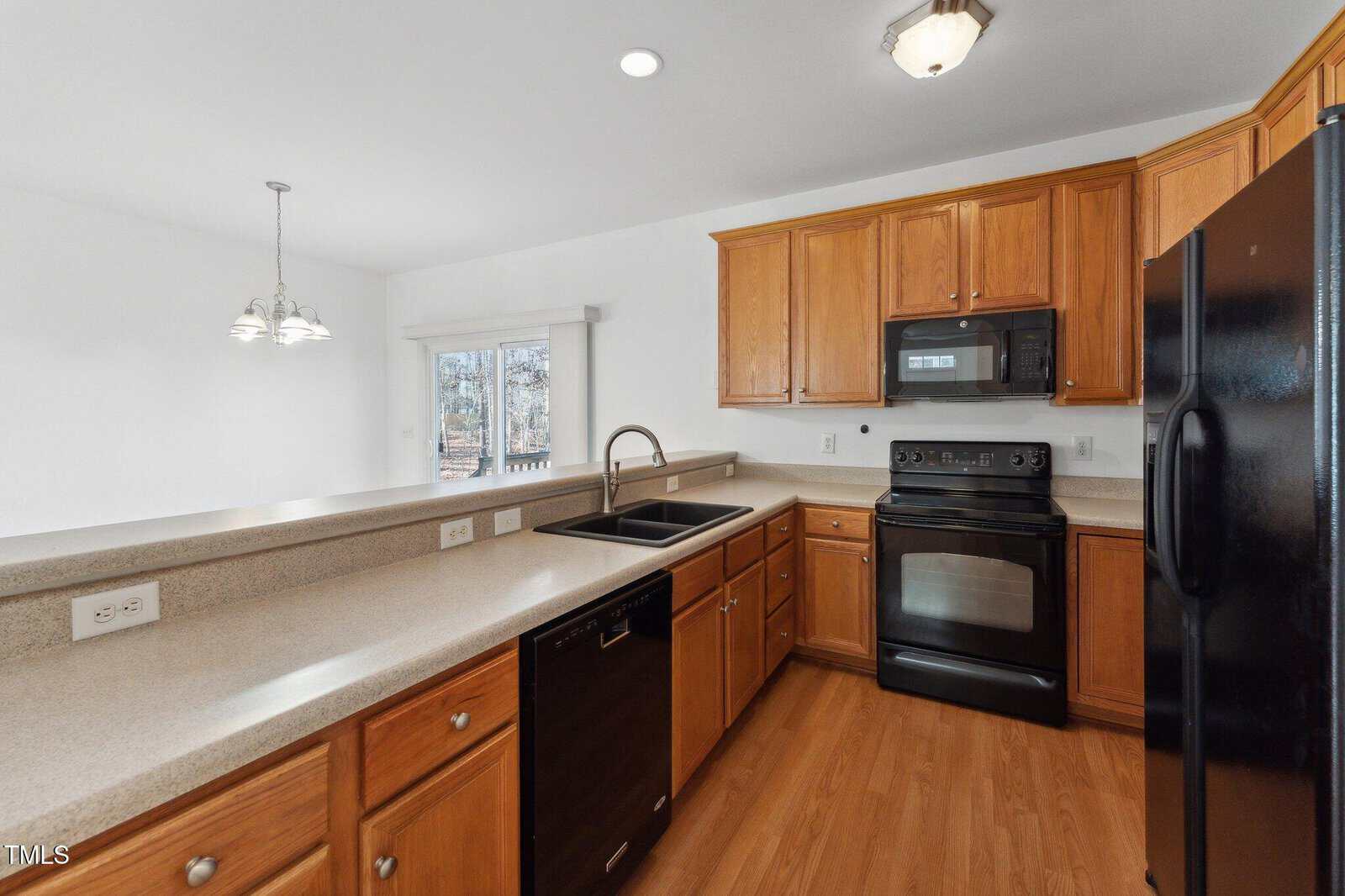 A kitchen with light wood cabinets, black appliances including a stove, microwave, refrigerator, and dishwasher, and a long beige countertop with a double sink. A dining area with windows is visible in the background.