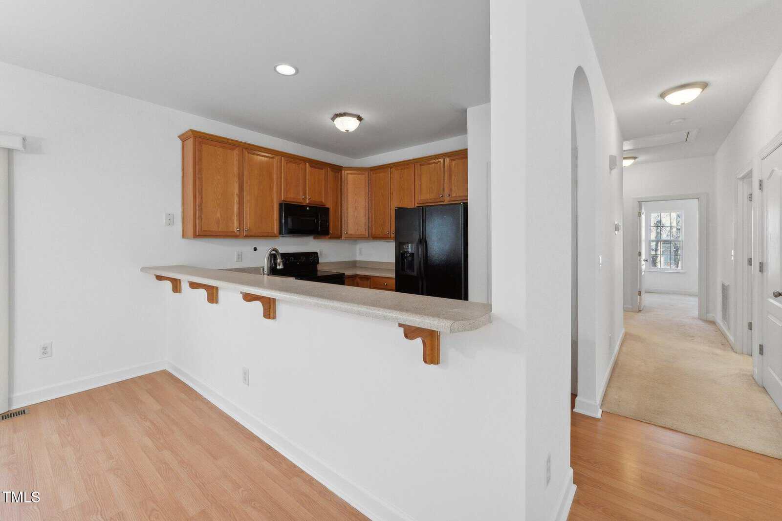 A kitchen with light wood cabinets, black appliances, and a long breakfast bar. Adjacent hallway leads to carpeted rooms and an arched doorway. Walls and ceiling are white.