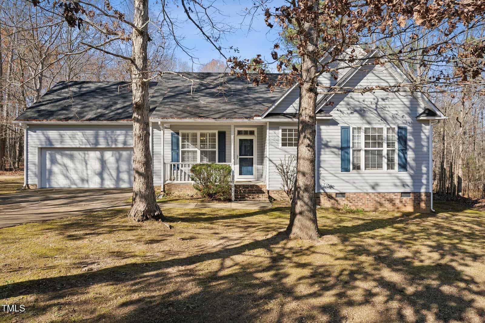A single-story light blue house with white trim, a two-car garage, front porch, and shutters. Leafless trees cast shadows on the front yard, and the sky is clear and blue.