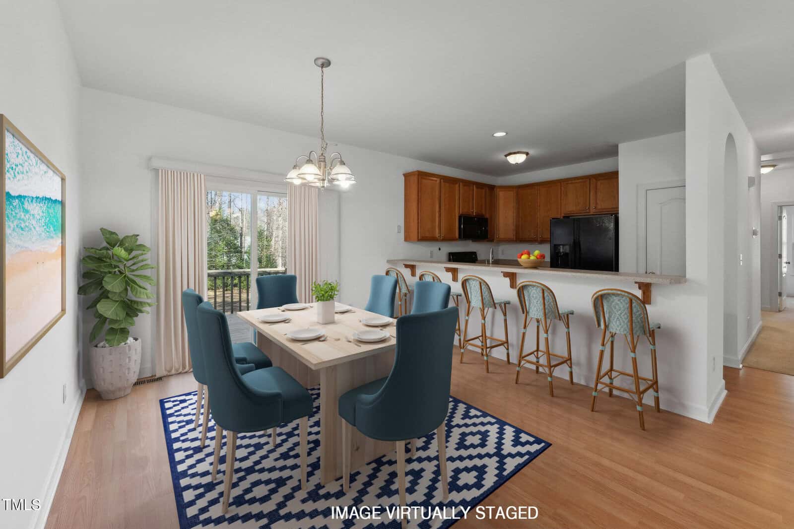 A bright dining and kitchen area with a square table, blue chairs, a patterned rug, barstools at the counter, wooden cabinets, and a large glass door overlooking trees. The image is virtually staged.