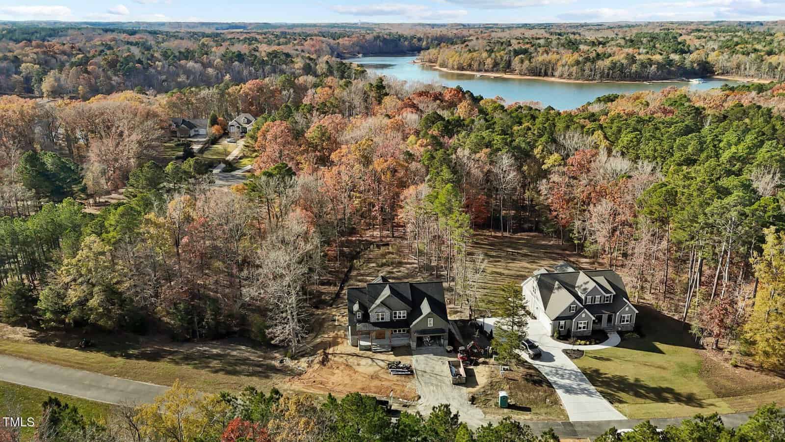 Aerial view of a wooded area with two large houses facing a quiet street. The landscape features autumn foliage and a serene lake in the background. The houses have spacious driveways and well-maintained lawns.