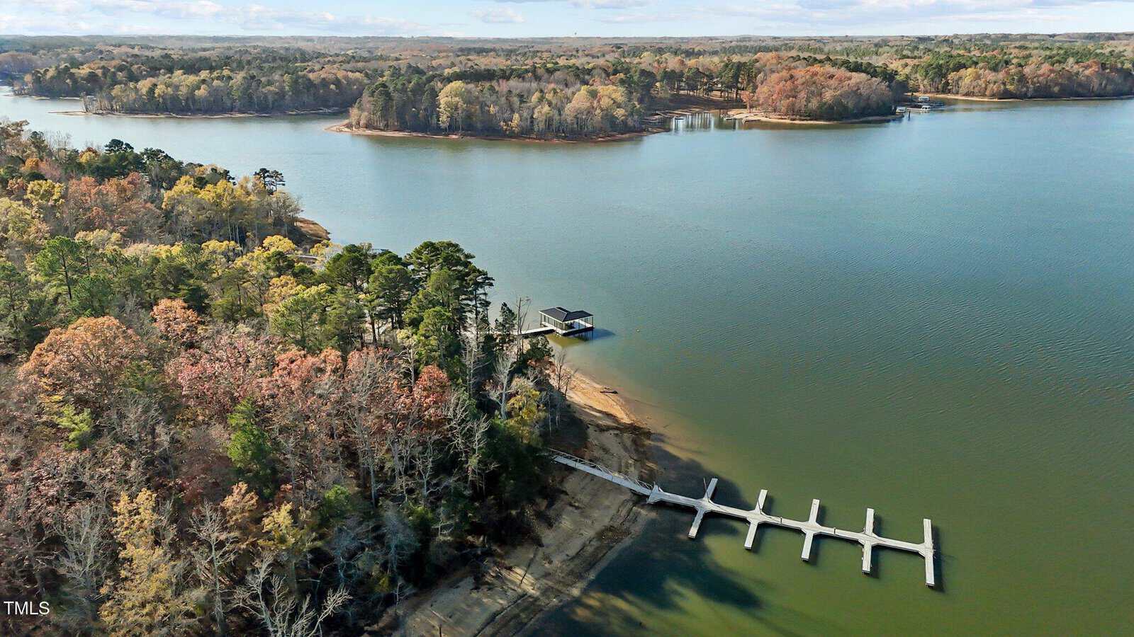 An aerial view of a calm lake surrounded by autumn trees. A dock extends into the water from a forested shoreline, with a small structure at the end. The distant shoreline is also lined with trees in fall colors under a partly cloudy sky.