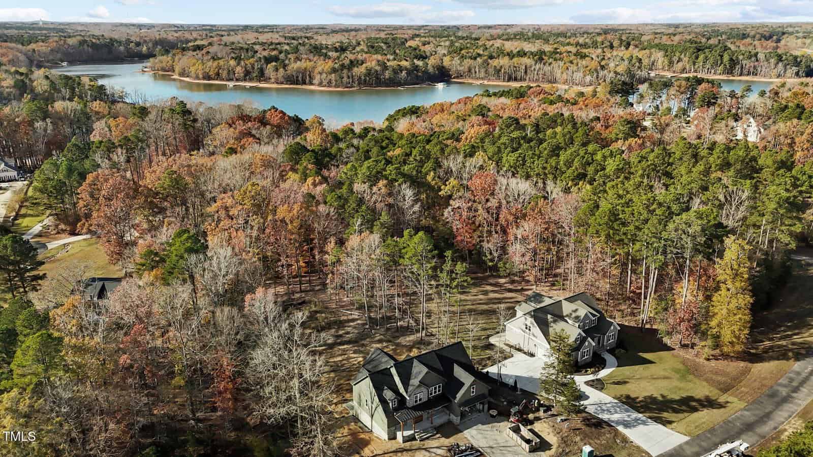 Aerial view of two houses surrounded by a forest of colorful autumn trees near a tranquil lake. The foliage is a mix of green, orange, and brown tones, and the water is calm under a partly cloudy sky.