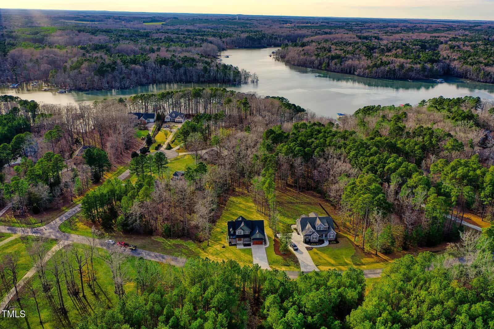 Aerial view of a suburban area with two large houses surrounded by trees. A winding road connects the neighborhood. A large lake is visible in the background, bordered by dense forest. The sky is partly cloudy.