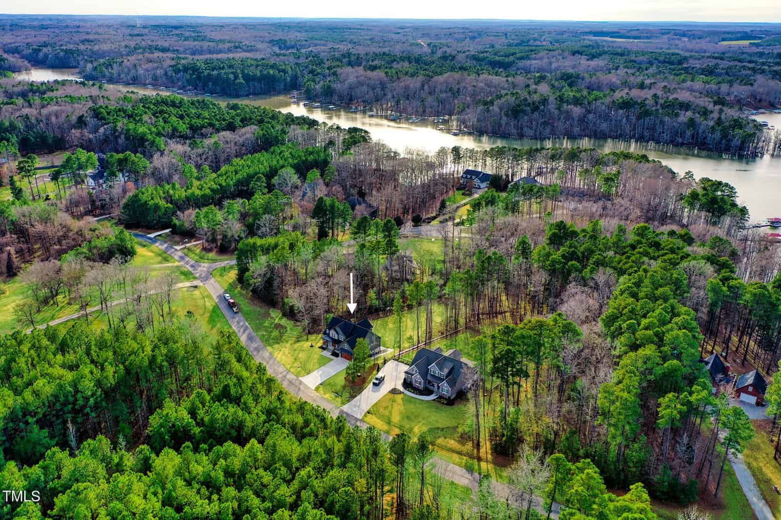 Aerial view of a residential area surrounded by green trees. Two houses are visible with driveways, and a river winds through the landscape in the background. An arrow points to one of the houses. The sky is partly cloudy.