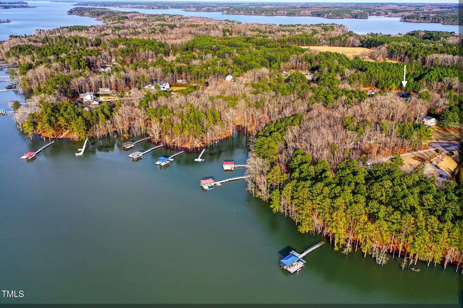 Aerial view of a lakeside landscape with dense green and brown forested areas. Several docks extend into the water, and scattered houses are visible among the trees. A large body of water surrounds the land, with more forested regions in the distance.