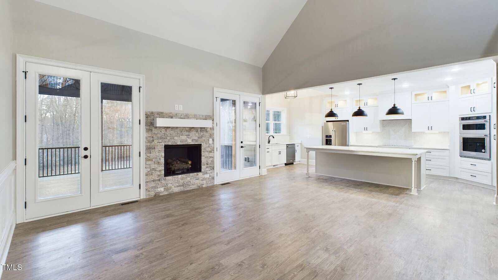 Spacious open-concept room with hardwood floors, featuring a kitchen with white cabinets and a large island with pendant lights. A stone fireplace is on the left with doors leading to a balcony. Large windows provide natural light.