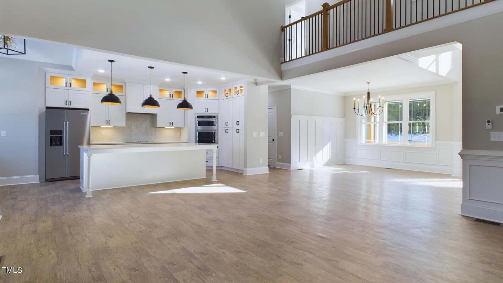 Spacious, modern open-plan kitchen and living area with light wood flooring. Features include a large island with white cabinets, three black pendant lights, stainless steel appliances, and a dining area with a window and chandelier.
