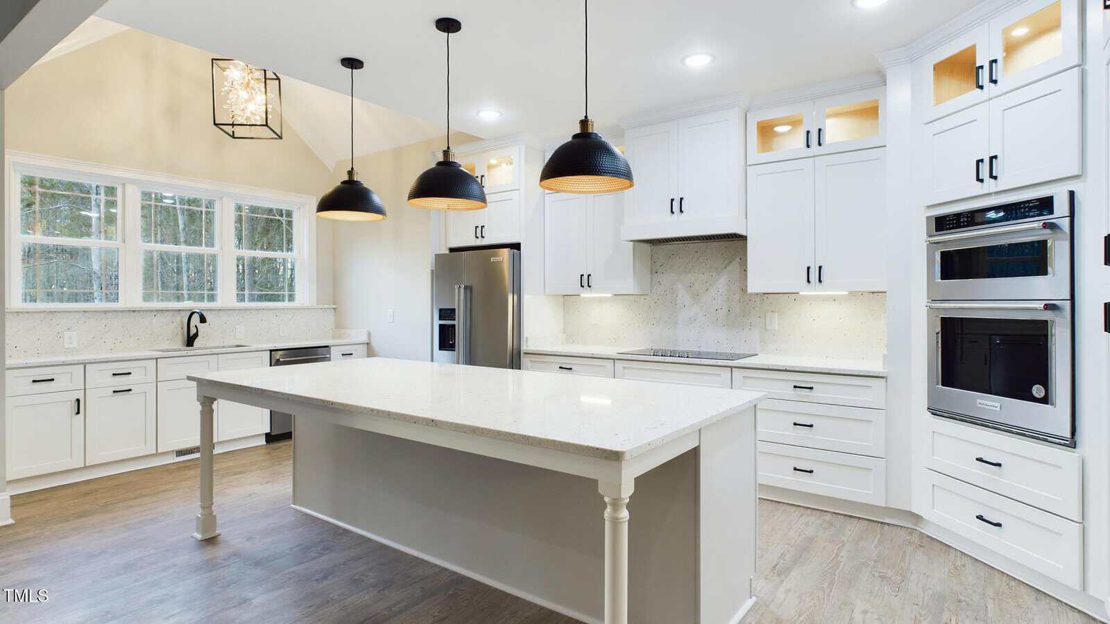 A modern kitchen with white cabinets and a large central island featuring a light countertop. Three black pendant lights hang above the island. Stainless steel appliances, including a fridge and double oven, are set against a backsplash.