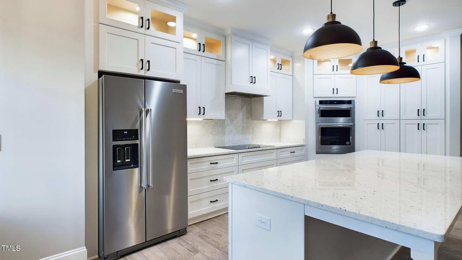 Modern kitchen with white cabinets, stainless steel refrigerator, built-in oven and microwave, and an electric cooktop. An island with a white quartz countertop and three pendant lights is in the foreground. The floor is light wood.