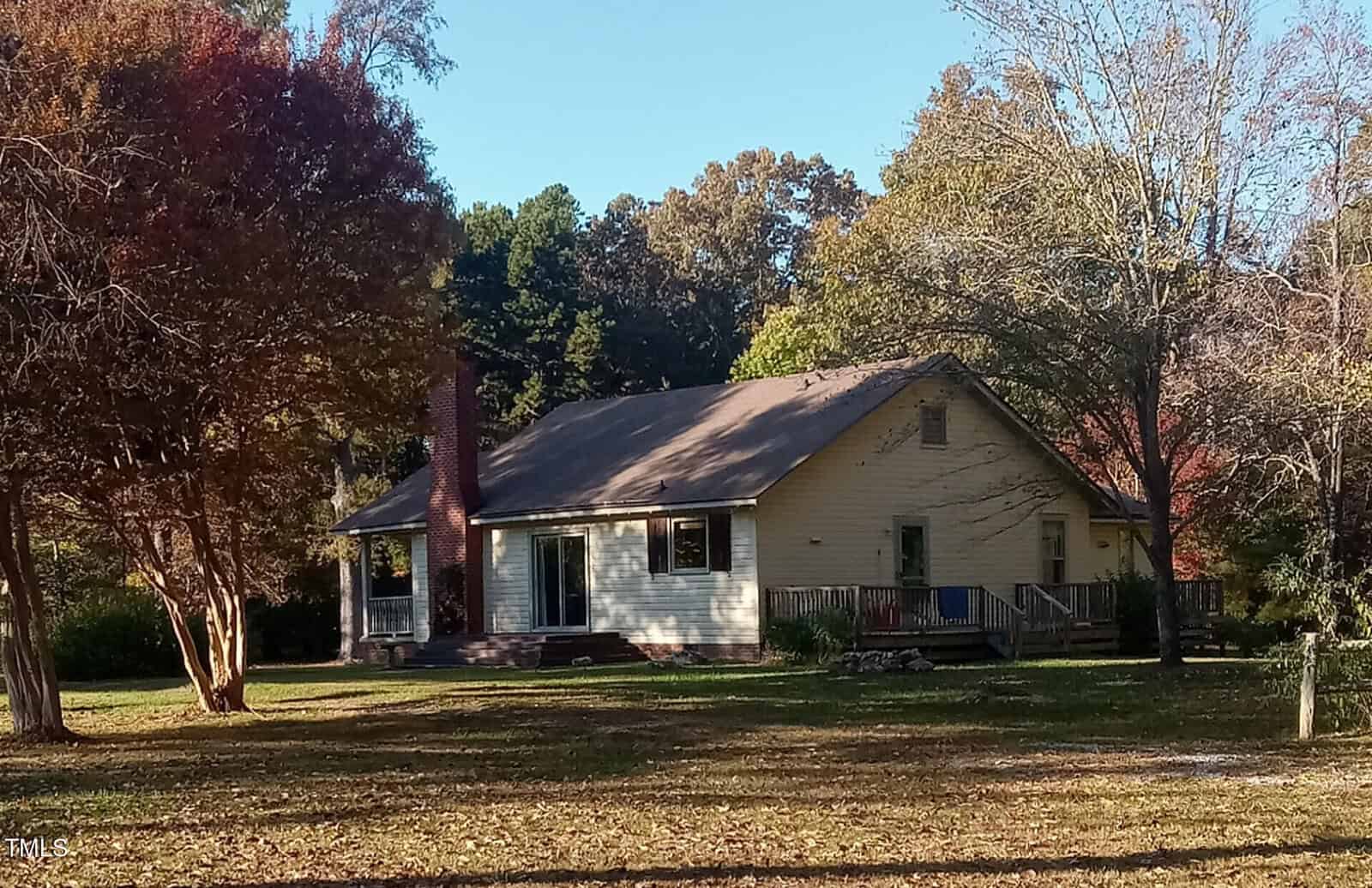 A single-story white house with a gable roof and a red brick chimney is surrounded by large trees. The house features a porch with railings and is set on a grassy lawn under a clear blue sky.