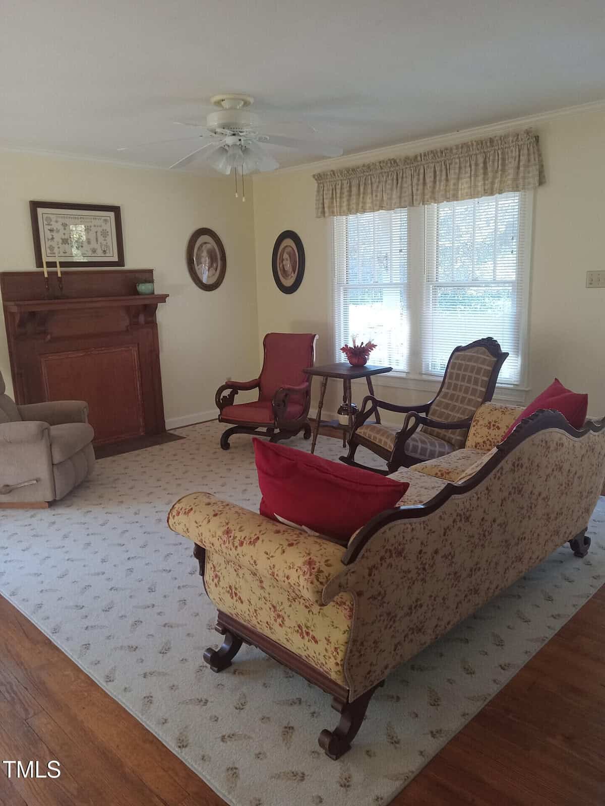 A living room with vintage decor features a patterned sofa, two armchairs, and a small wooden table. A fireplace is against the far wall, and a window with curtains lets in natural light. The room has a ceiling fan and hardwood floors partly covered by a rug.