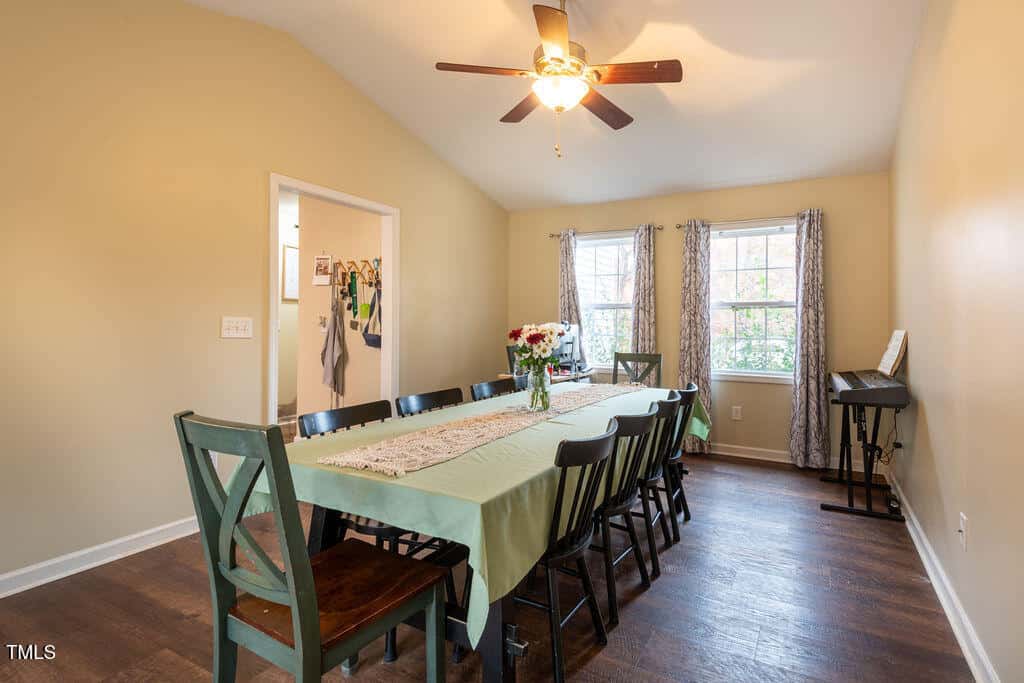 A dining room with a long table covered by a green tablecloth, black chairs, flowers in a vase, hardwood floor, ceiling fan, two windows with curtains, and a keyboard in the corner.