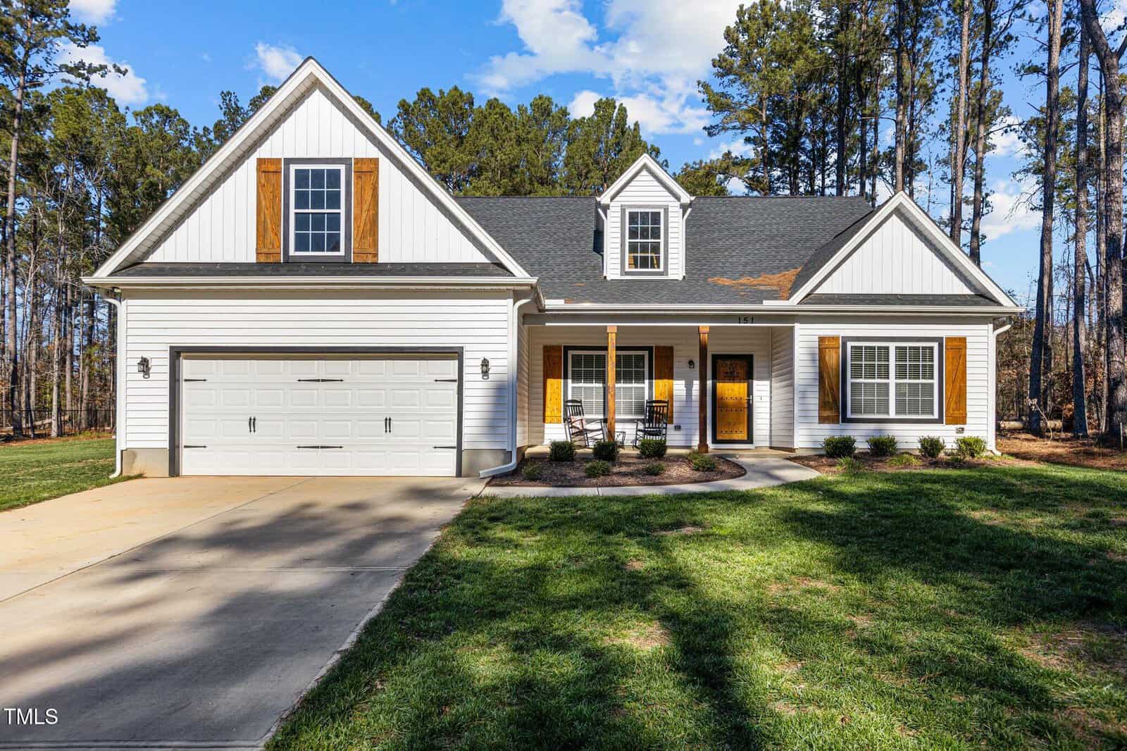 A single-story house with white siding, wood accents, a two-car garage, a covered front porch, and a well-kept lawn, surrounded by tall trees under a partly cloudy sky.