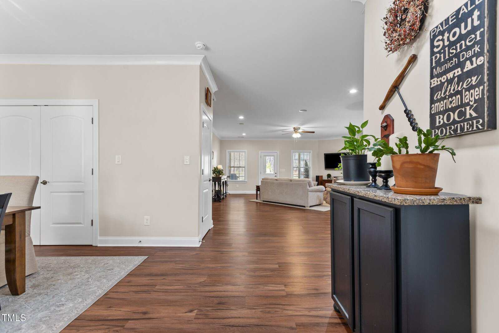 A hallway with wood flooring leads to a living area with a sofa, armchair, and TV. A console table with potted plants and wall decor is on the right; double doors and a dining area are partially visible on the left.