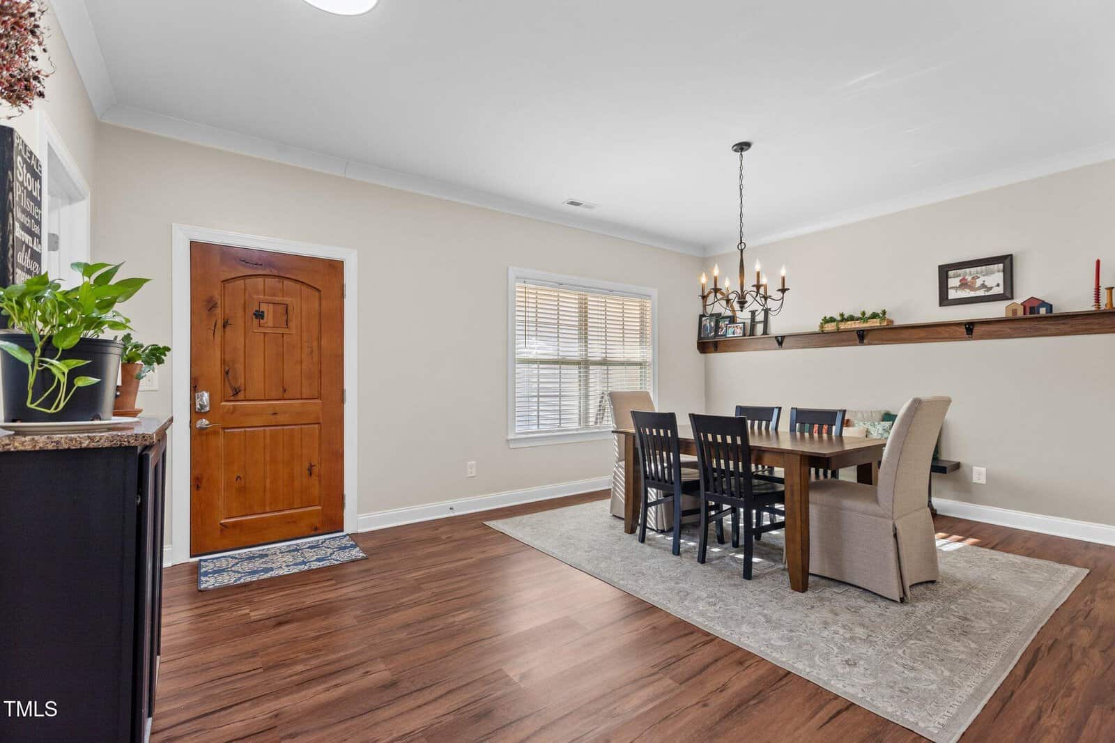 Dining area with a wooden table, six chairs, and a beige rug. A wooden door and window are on one wall; plants, a sideboard, and a long shelf with dรฉcor accent the neutral, cozy space.
