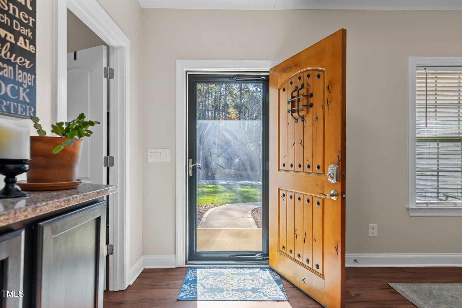 A wooden front door with metal accents is open, revealing a glass storm door. Sunlight streams in, illuminating a blue rug. Part of a kitchen counter, a white wall, and a window with blinds are visible.