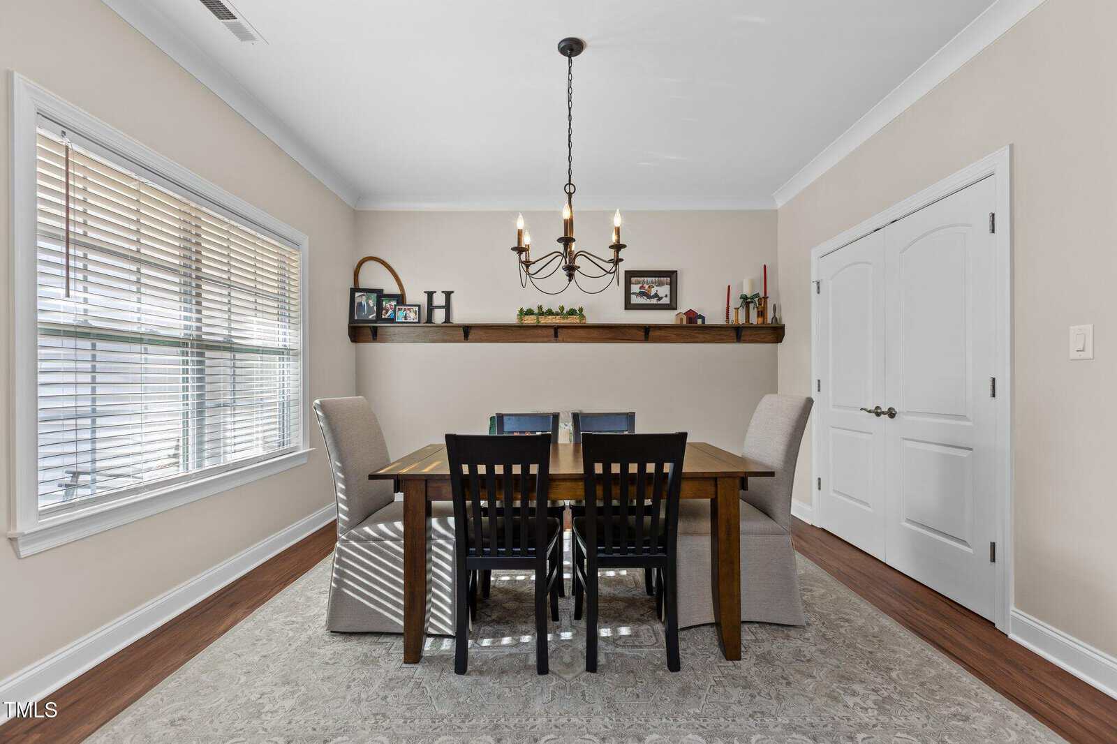 A dining room with a wooden table, four black chairs, two beige armchairs, a chandelier, a window with blinds, a rug, and a shelf displaying decor items on a beige wall.