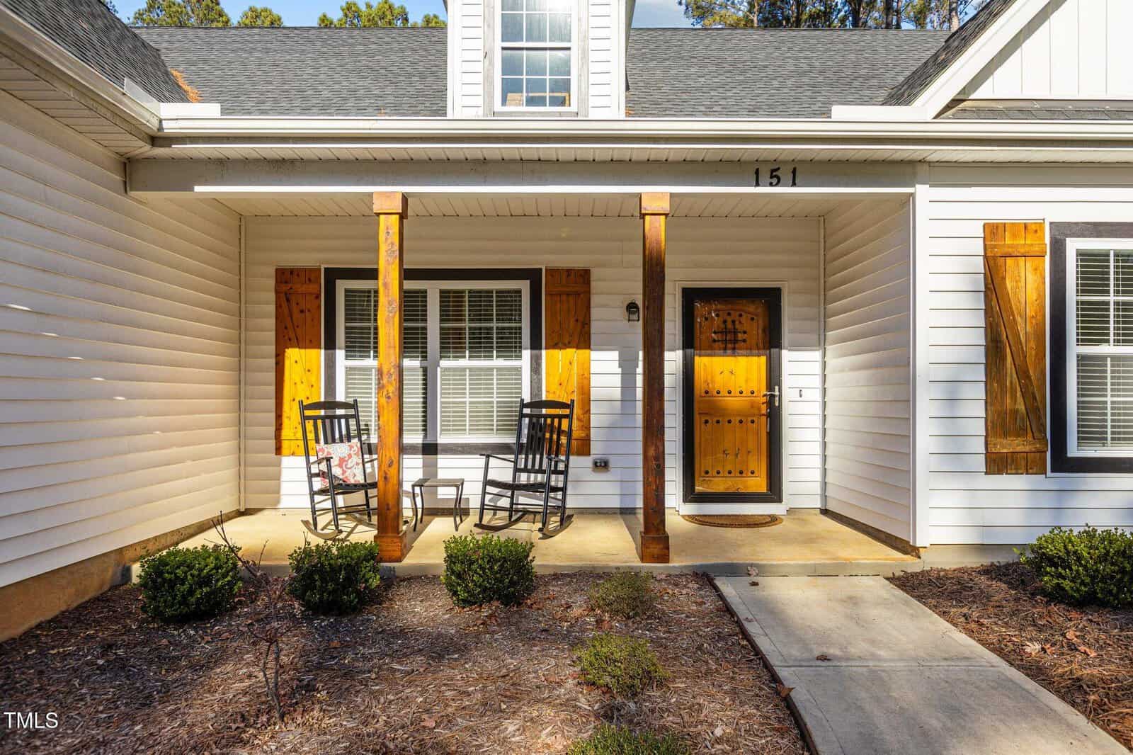 A white house exterior with wood accents, featuring a covered porch with two black rocking chairs, a wooden door, and a concrete walkway leading to the entrance. Small shrubs line the front of the porch.