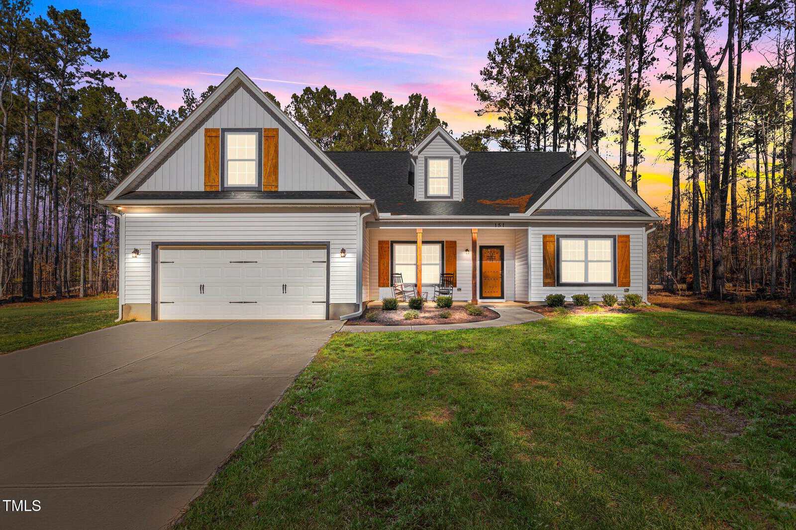A single-story white house with wooden shutters, a double garage, and a front porch sits on a grassy yard with a concrete driveway. Tall trees and a colorful sunset sky are in the background.