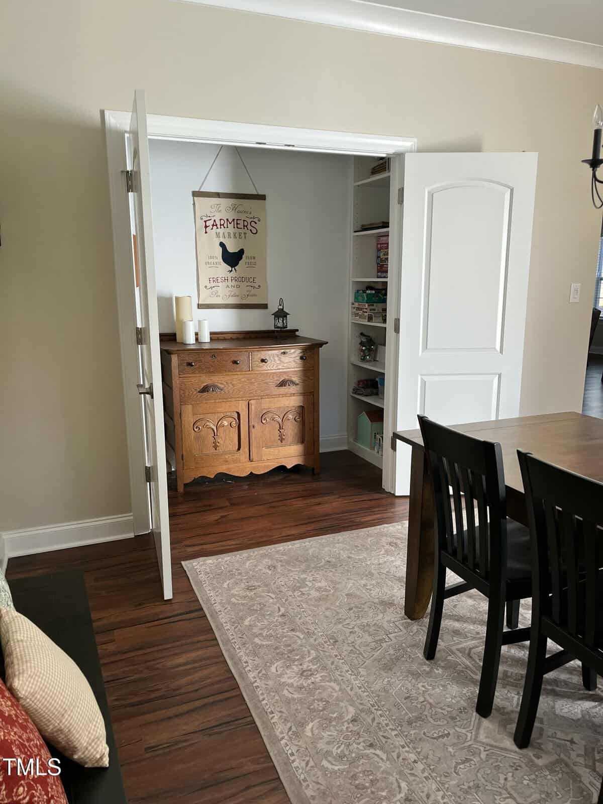 A dining room with dark wood table and chairs, a light patterned rug, and open double doors revealing shelves and a small wooden cabinet with a decorative farm-themed sign above it.