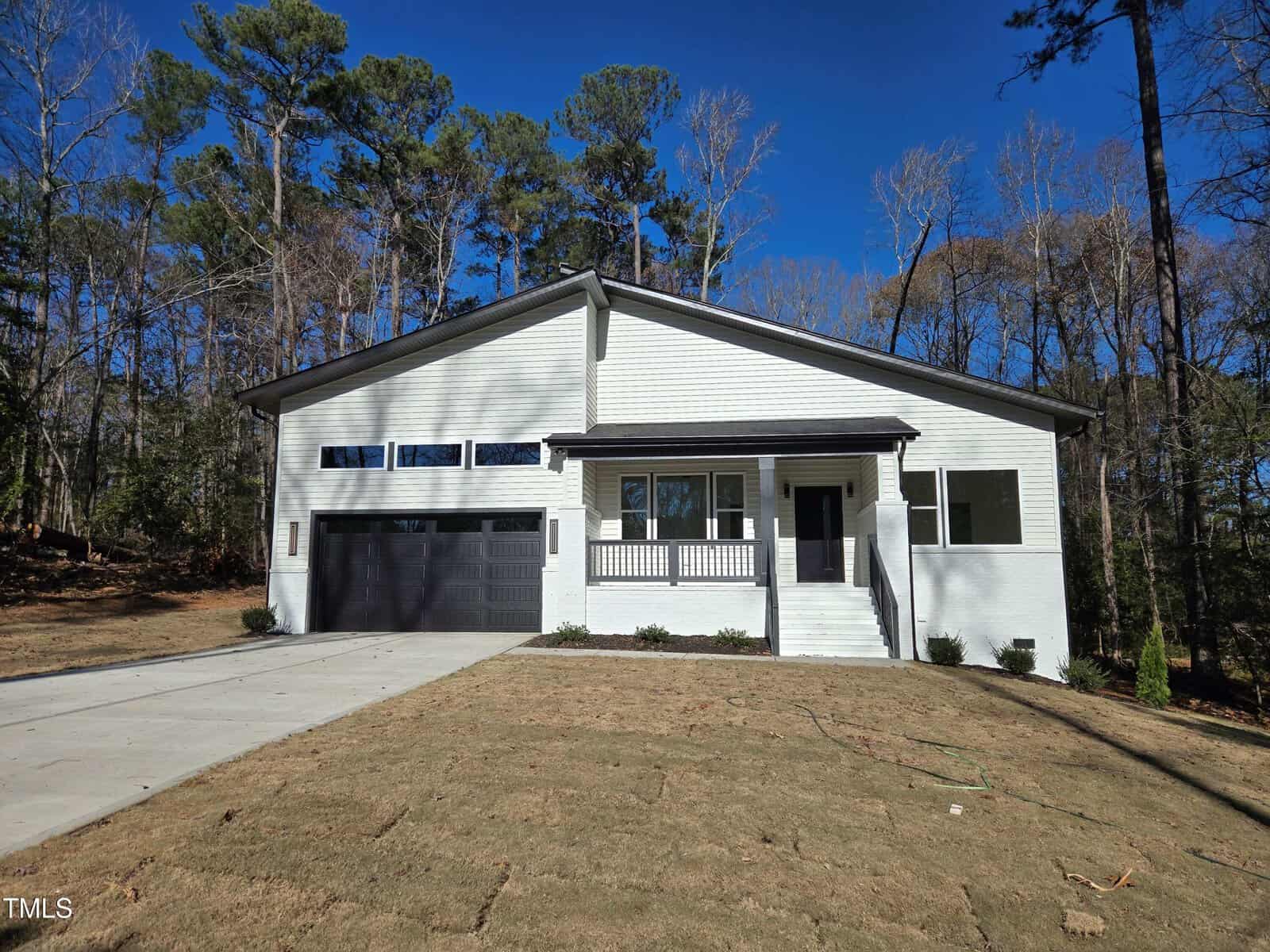A modern single-story house with white brick exterior, black trim, and a black double garage. The home has a front porch with steps and sits on a newly sodded lawn surrounded by tall trees.