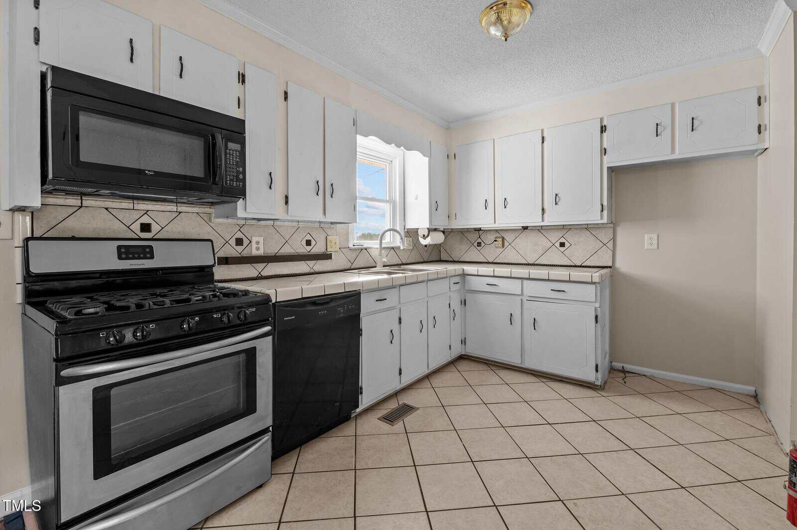 Kitchen with white cabinets, a black dishwasher, and a black oven with a gas stovetop. The walls have light tile backsplashes with diamond patterns. A window lets in natural light, and the floor is covered with beige tiles.