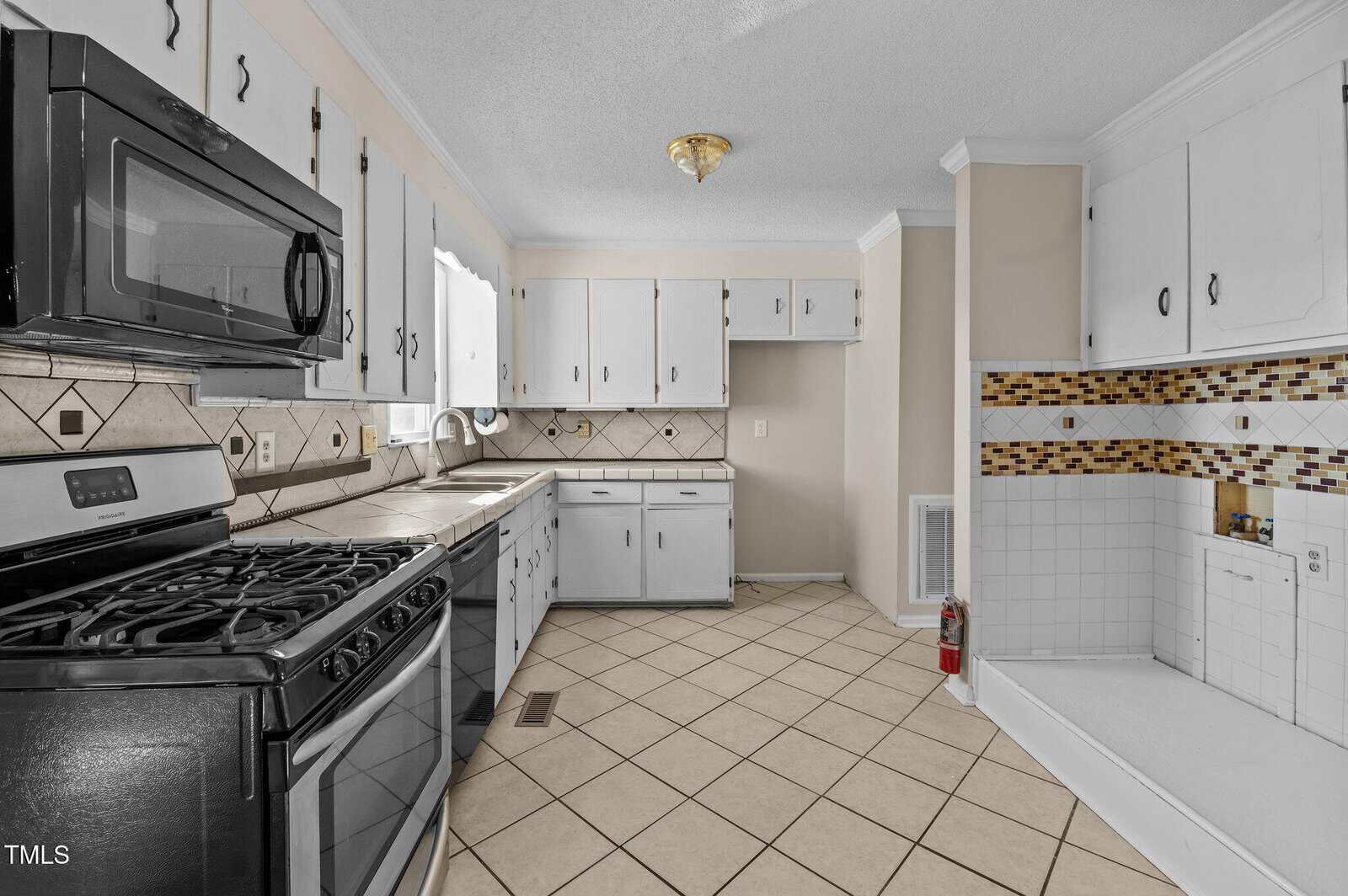 A kitchen with black appliances, including a stove and microwave. White cabinets contrast with a tiled backsplash, featuring a mix of diamond and mosaic patterns. Beige walls and a tiled floor complement the room. A fire extinguisher is on the floor.