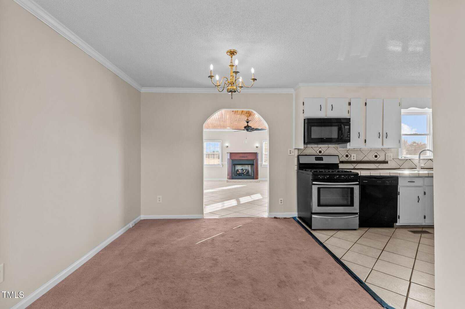 An empty kitchen and dining area with beige walls and carpet, featuring a chandelier. The kitchen includes a black stove, microwave, and tiled backsplash. An arched opening leads to a sunlit room with a fireplace and ceiling fan in the background.