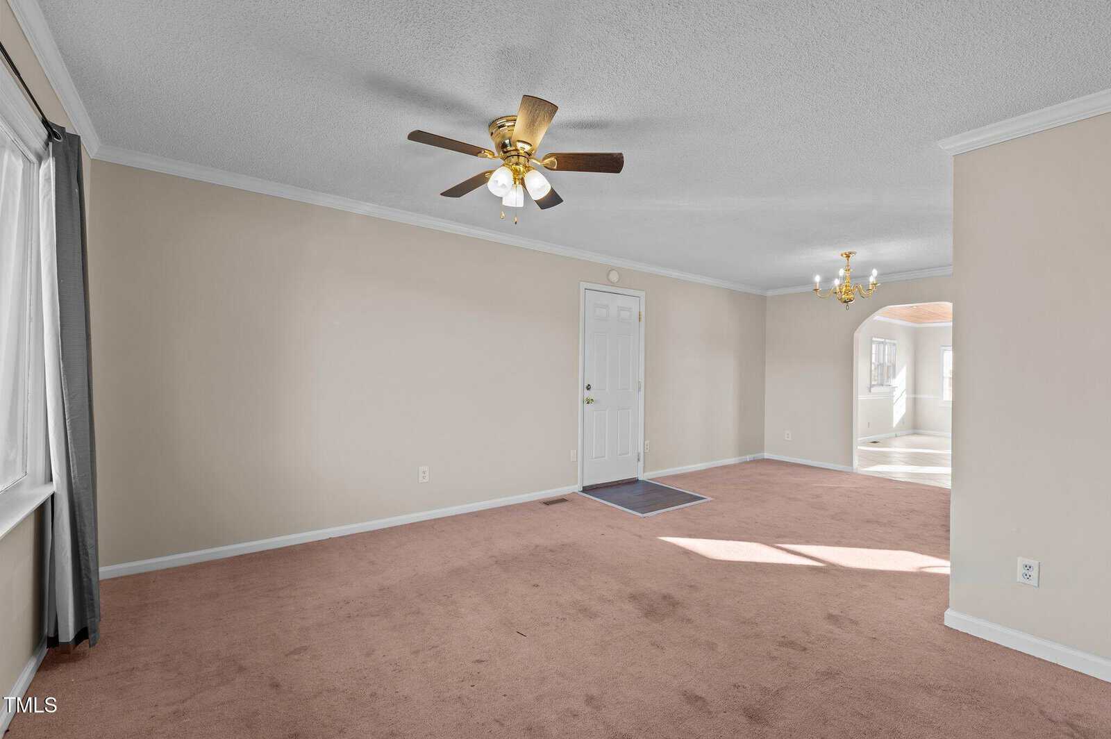 A spacious, empty living room with beige carpet, light gray walls, a ceiling fan, and a chandelier. The room has a large window with dark curtains and a view into a sunlit adjacent room through an open doorway.
