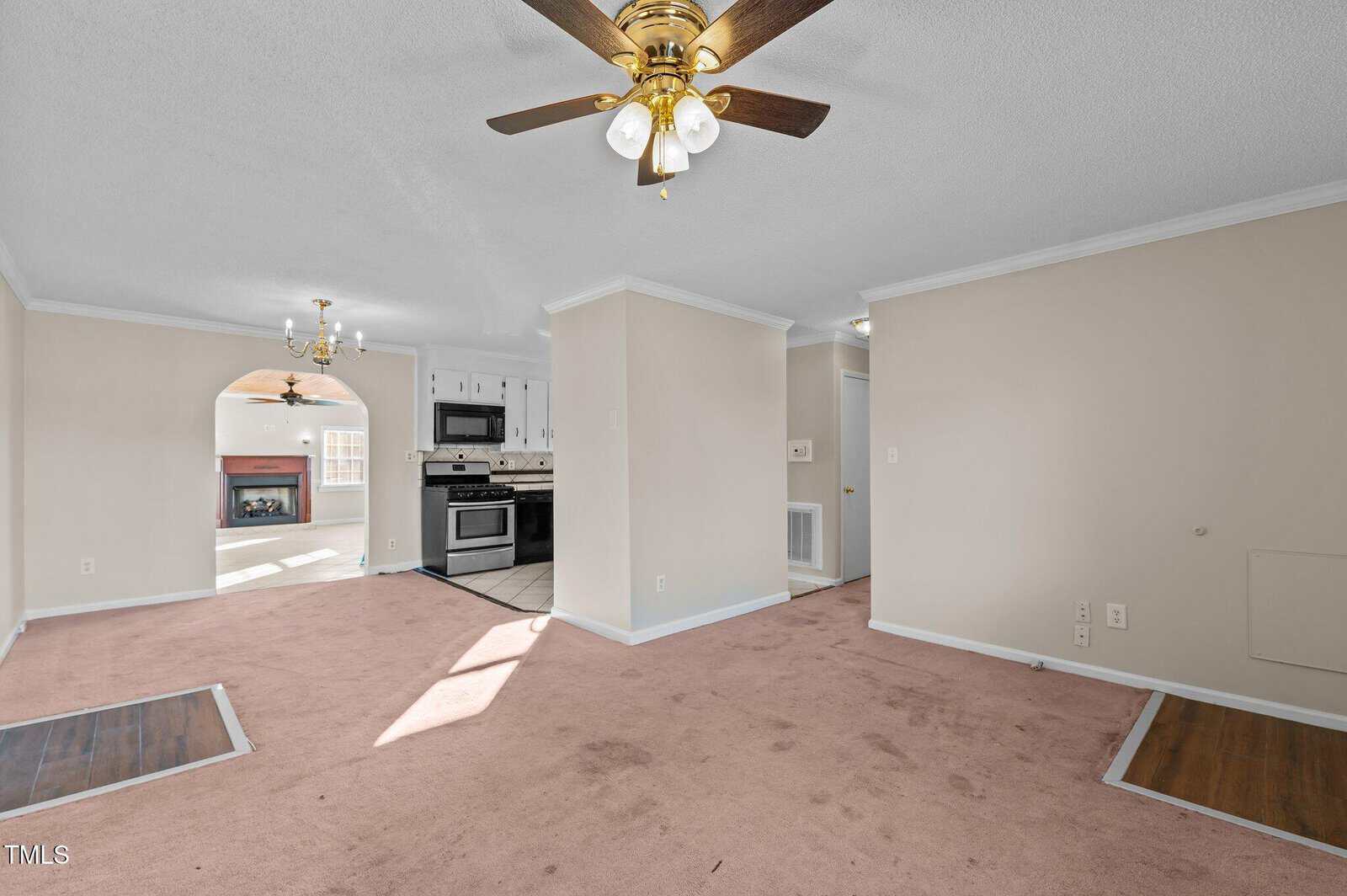 A spacious room with beige walls and carpet features two ceiling fans with lights. The area includes a view of a kitchen with white cabinets and a dining nook with a chandelier. Sunlight streams through the windows, illuminating the space.