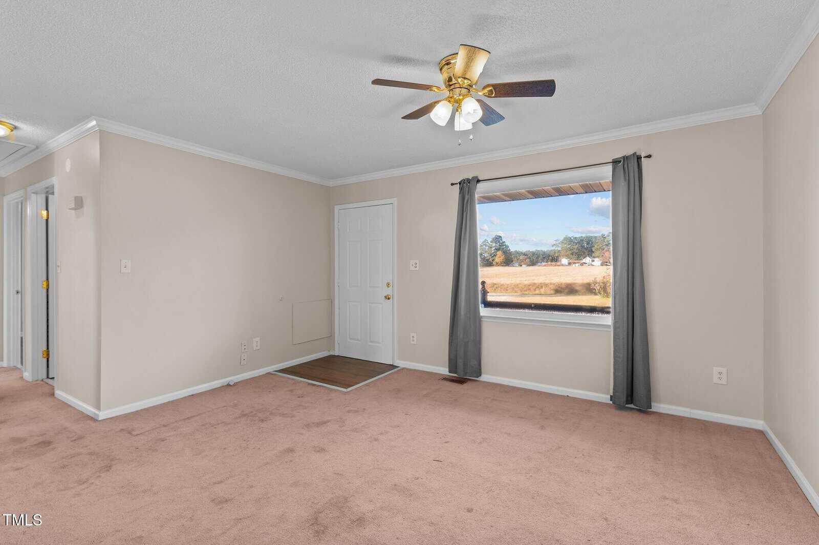 A bright, empty room with beige carpet and light-colored walls. A ceiling fan is centered on the white ceiling. There's a large window with gray curtains, and a white door near the corner. Hallways lead to other rooms.