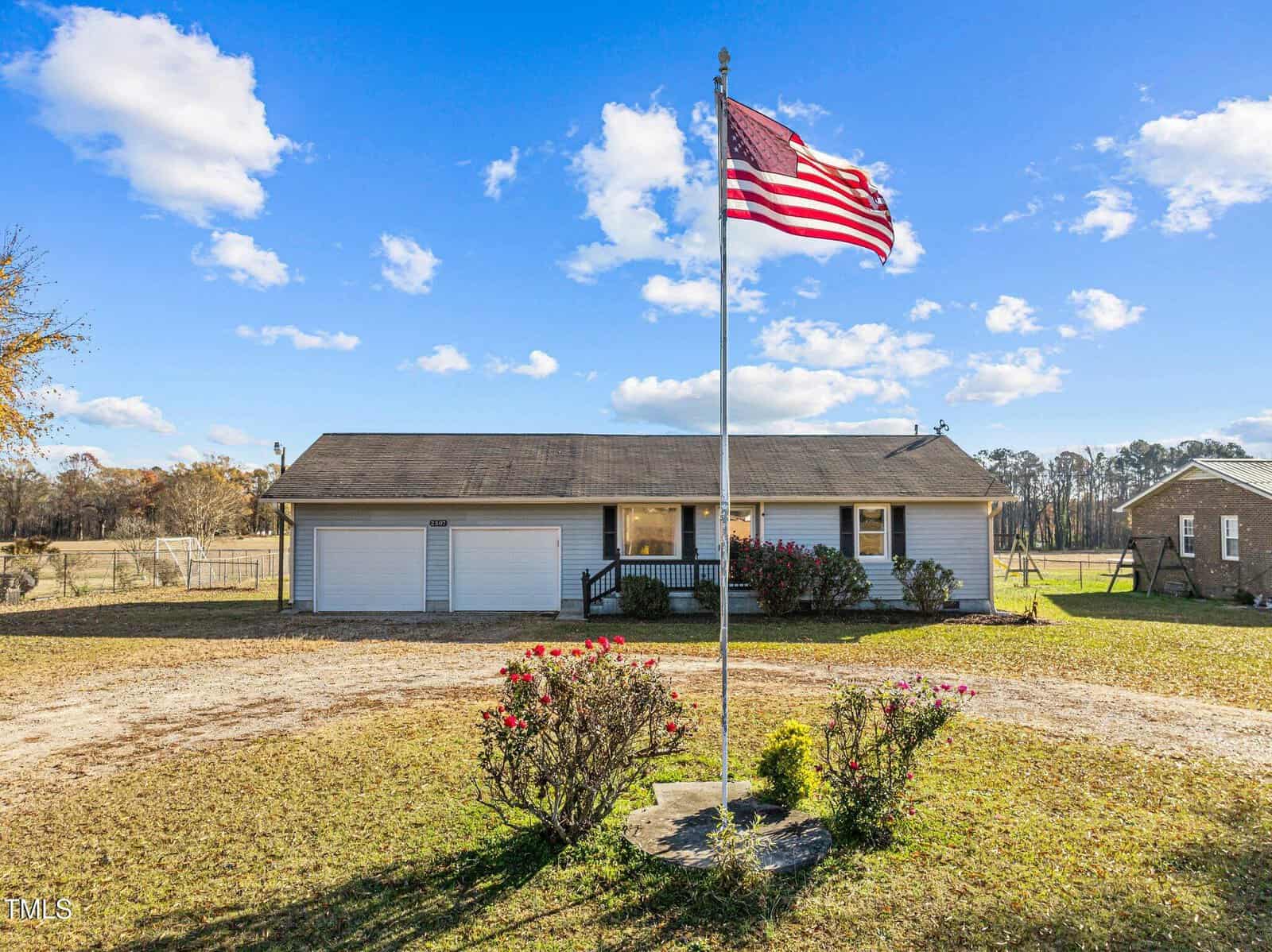 A single-story house at 2507 Rock Mill Road features a welcoming front porch and a spacious driveway. An American flag proudly waves in front, complementing the yard's shrubs and grass. The sky overhead is clear with a few clouds, creating a charming Henderson setting.