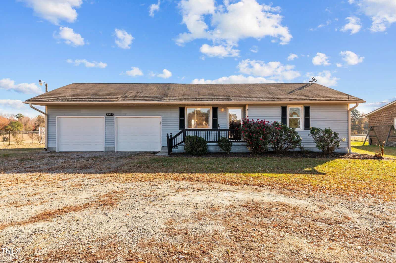 A single-story house with two white garage doors, a small front porch, and a sloped roof. The yard has some grass and a gravel driveway. The sky is bright with scattered white clouds.