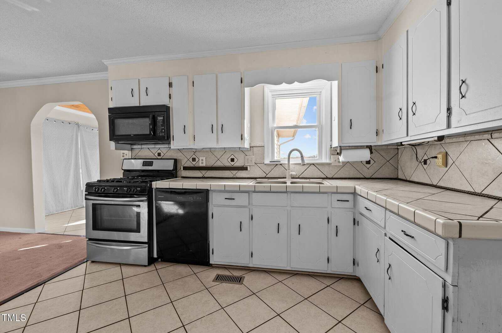 A kitchen with white cabinets and tiled countertops, featuring a black oven, microwave, and dishwasher. A single window provides natural light, and the floor is covered in beige tiles. An arched doorway leads to another room.