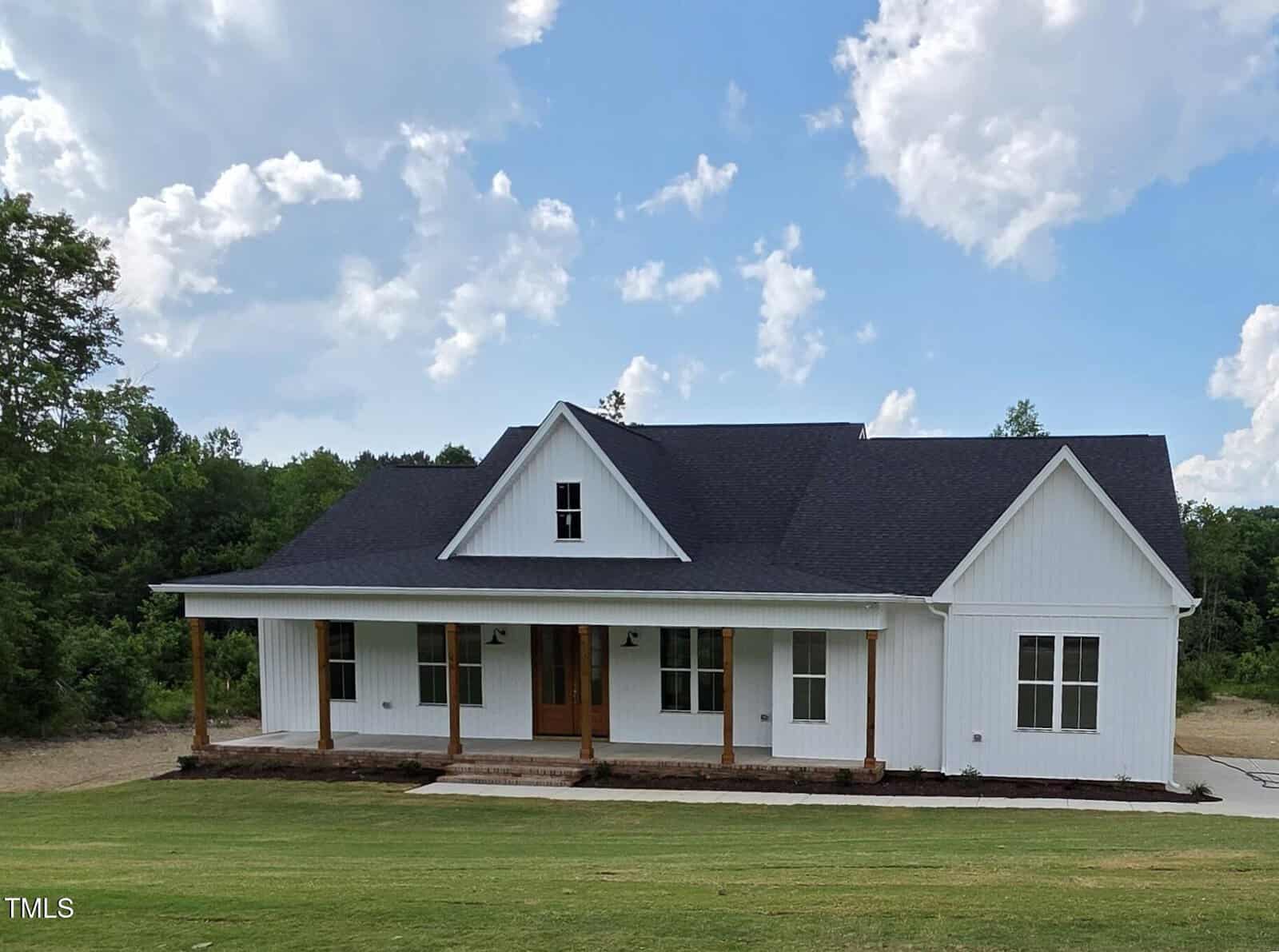 A white, single-story farmhouse with a black roof, wooden porch columns, and a covered front porch sits on a grassy lawn with trees in the background under a blue sky with scattered clouds.
