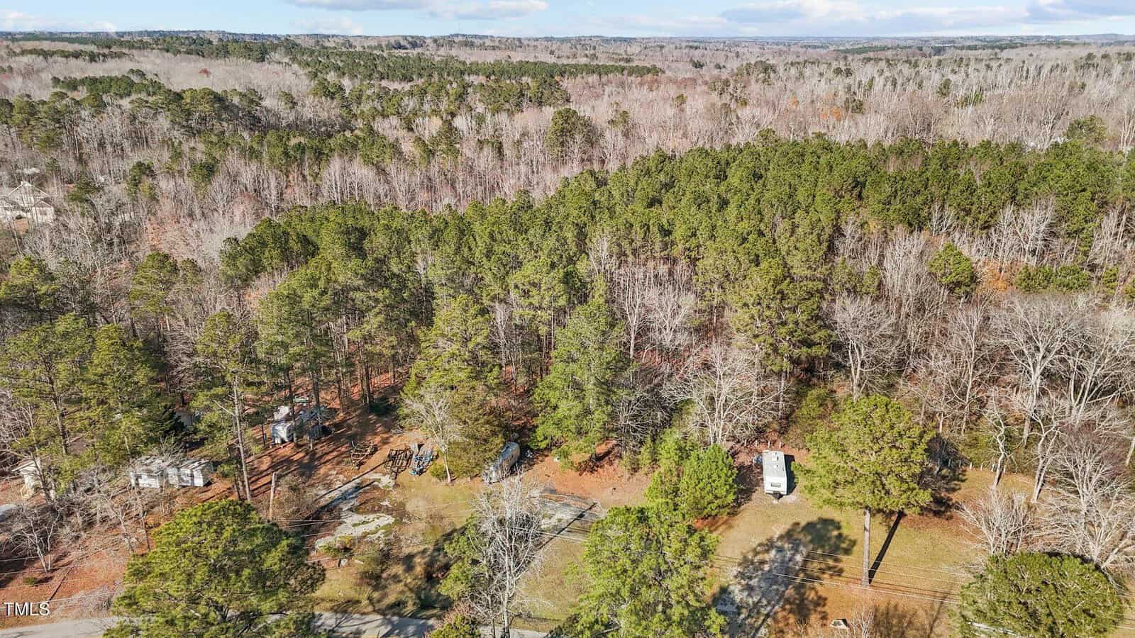 Aerial view of a wooded rural area with dense trees, some open clearings, a few buildings or structures, a white vehicle, and a road running through the lower part of the image. Hills and forest extend into the distance.