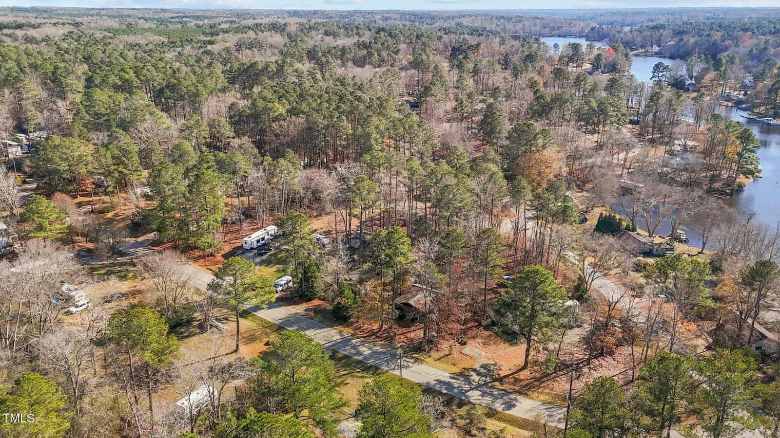 Aerial view of a wooded lakeside area with scattered houses, RVs, and a winding road. The lake curves through the trees, and some trees have lost leaves, suggesting a late autumn or early winter setting.