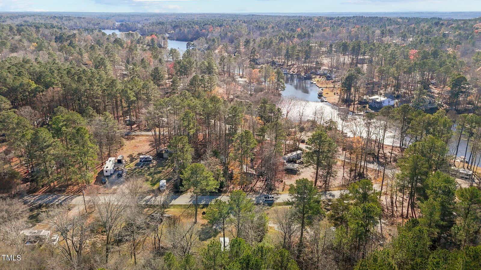 Aerial view of a wooded area with scattered houses, RVs, and a winding lake. Trees with autumn leaves surround the water, and a road runs through the scene. The sky is partly cloudy.