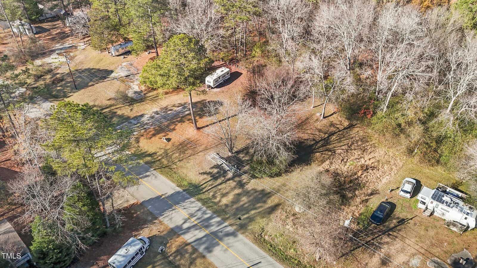 Aerial view of a rural road with several RVs and vehicles parked along the roadside and under trees, bordered by wooded and grassy areas with leafless trees.