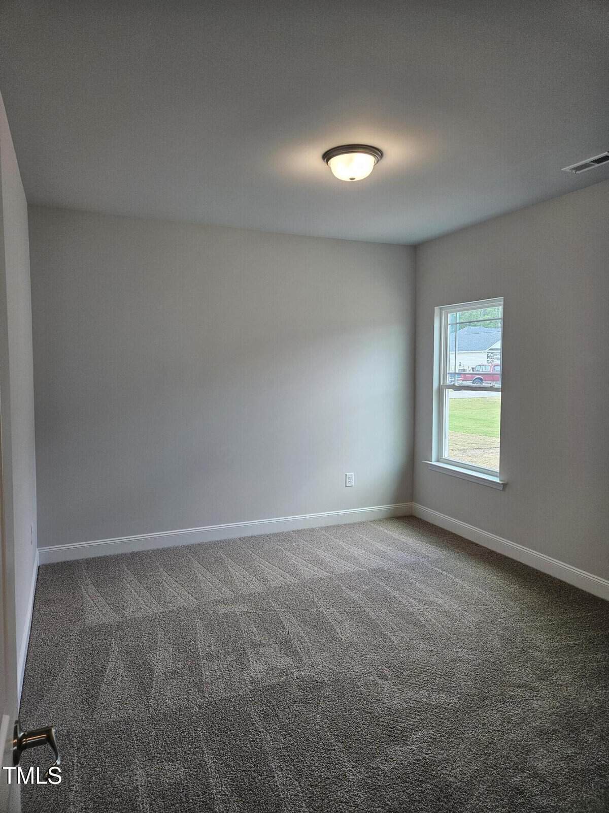 Empty carpeted room with light gray walls, a ceiling light, and one window letting in natural daylight. A small portion of a neighborhood can be seen outside through the window.