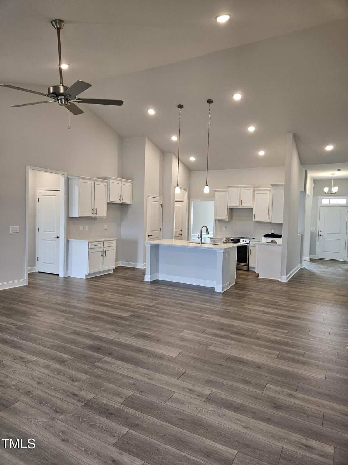 Open-concept kitchen and living area with white cabinets, pendant lights over an island, stainless steel appliances, and light gray walls. Wood-style flooring extends throughout the space. Ceiling fan and recessed lighting are visible.