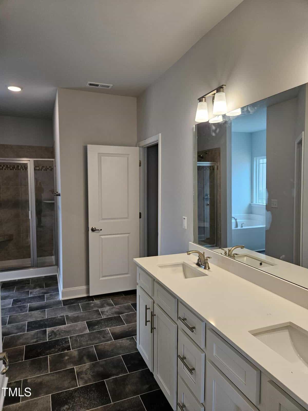 Modern bathroom with double sink vanity, large mirror, dark tile floor, glass shower enclosure, white cabinets, and a separate bathtub visible through the doorway.
