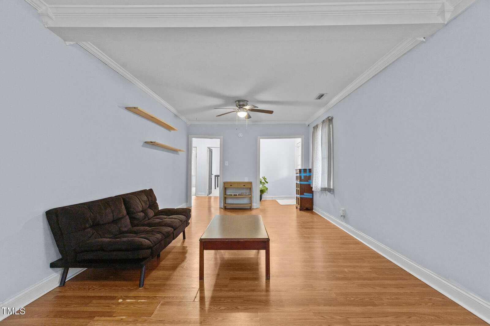 A living room with light blue walls and wooden flooring features a black futon on the left, a wooden coffee table in the center, and wall-mounted shelves. A ceiling fan is overhead, and windows with blinds allow natural light. A doorway is at the far end.