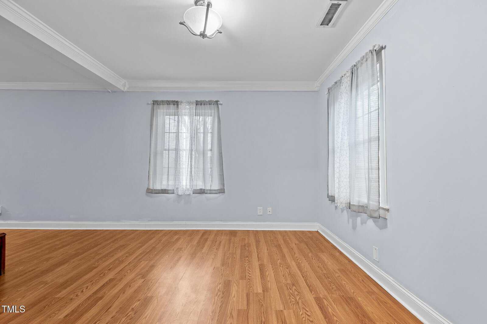 An empty room with light blue walls and wooden flooring. It features two windows with sheer curtains, a white ceiling with crown molding, a modern ceiling light fixture, and two power outlets on one wall.