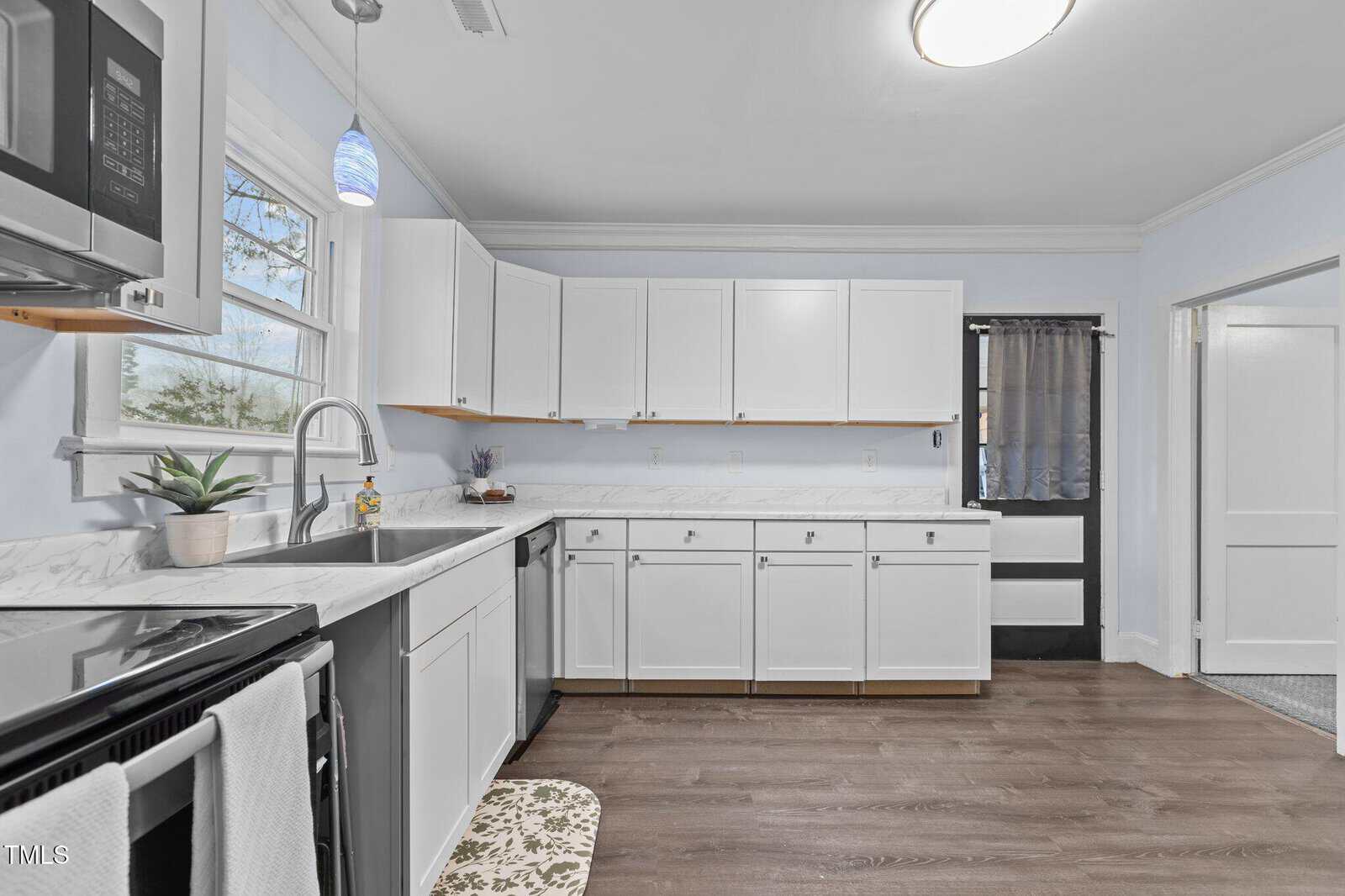 A modern kitchen with white cabinets, a stainless steel oven, and a built-in microwave. There is a potted plant on the countertop, a blue pendant light above, and a window near the sink. The floor is made of wood, and the walls are painted light blue.