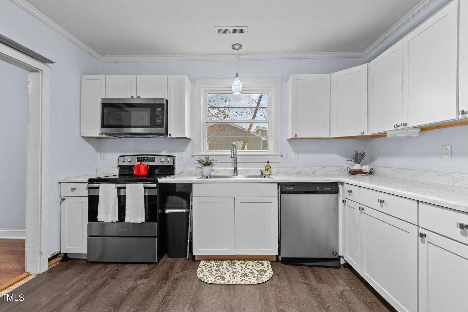A modern kitchen with white cabinets and marble countertops. There is a stainless steel oven with a microwave above it, a dishwasher, and a window with a view of trees. A pendant light hangs from the ceiling, and a small indoor plant is on the counter.