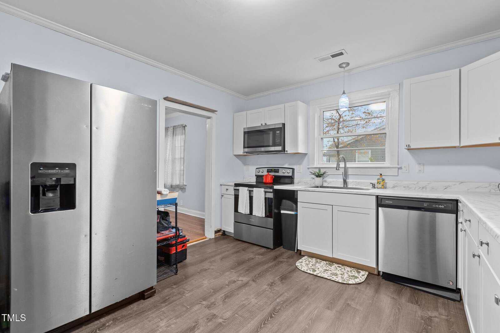A modern kitchen with stainless steel appliances, including a refrigerator, oven, and dishwasher. White cabinets and a marble-patterned countertop complement the light blue walls. A window above the sink lets in natural light.