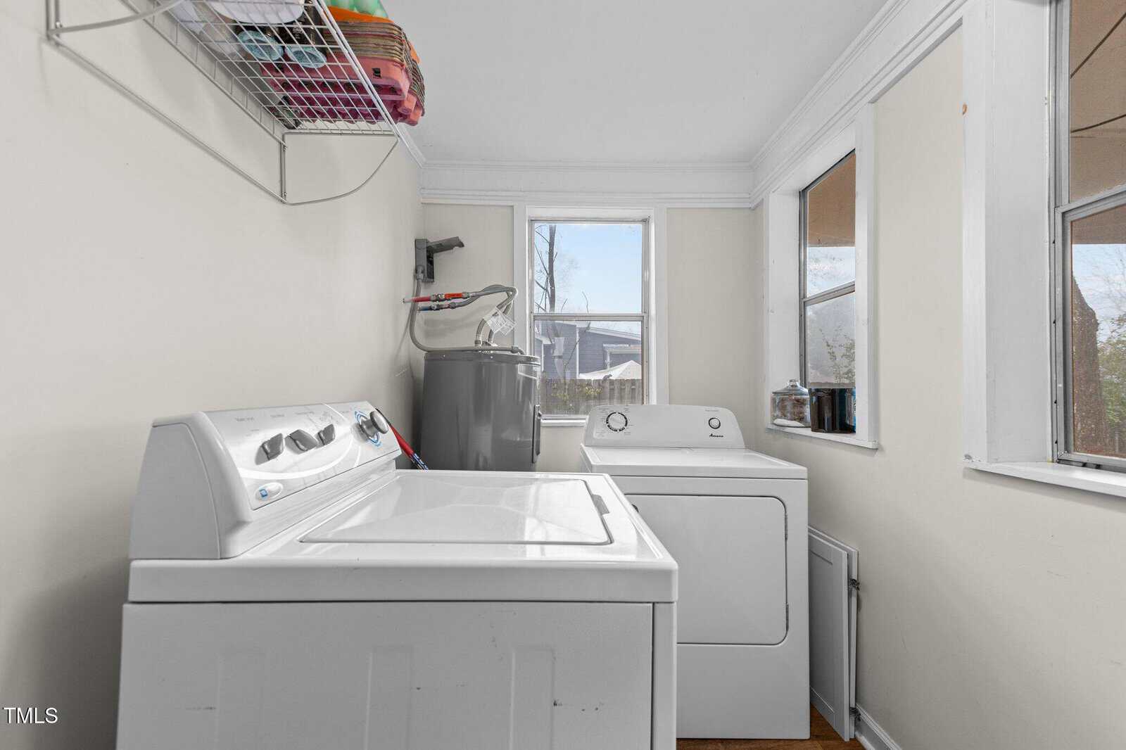 Laundry room with a white washing machine and dryer side by side against a pale wall. Above them is a wire shelf holding colorful towels. The room has a window to the right and a larger window ahead, both letting in natural light.